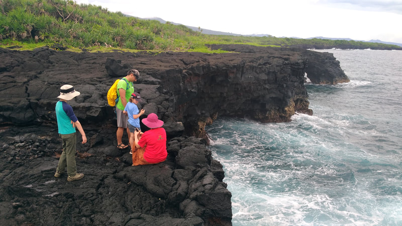 A family in colourful clothing sit on the lava fields and admire the ocean view on the Coastal Cliff Walk, a part of Samoa's O le Pupu-Pue National Park.